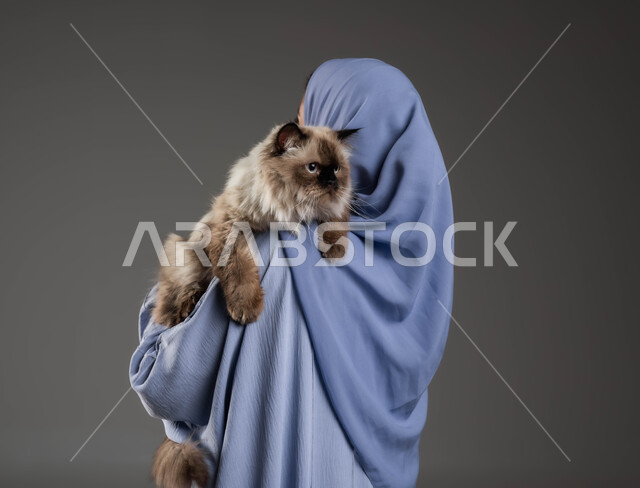 الرفق بالحيوان والعطف عليه، الأثر الايجابي لاقتناء الحيوانات المنزلية وتربيتها، بورتريه مقرب من الخلف لإمرأة عربية خليجية سعودية ترتدي الحجاب والعباءة تقوم بحمل قطة رمادية أليفة، الشعور بالسعادة أثناء قضاء وقت ممتع في اللعب مع القط، خلفية رمادية