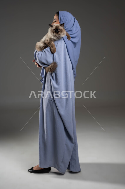 الرفق بالحيوان والعطف عليه، الأثر الايجابي لاقتناء الحيوانات المنزلية وتربيتها، بورتريه من الخلف لإمرأة عربية خليجية سعودية ترتدي الحجاب والعباءة تقوم بحمل قطة رمادية أليفة، الشعور بالسعادة أثناء قضاء وقت ممتع في اللعب مع القط، صورة لكامل الجسم، خلفية رمادية