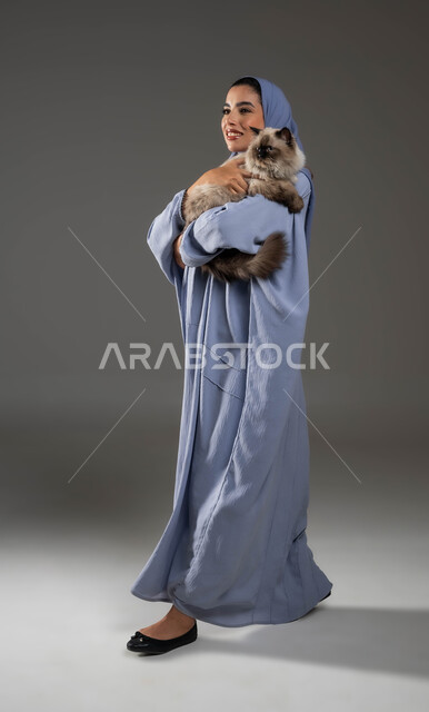 ايماءات المشي والذهاب إلى مكان ما، بورتريه لإمرأة عربية خليجية سعودية مبتسمة ترتدي الحجاب والعباءة تقوم بحمل قطة رمادية، قضاء وقت ممتع في اللعب مع القط، مفهوم الرفق بالحيوان، الأثر الايجابي لاقتناء الحيوانات المنزلية وتربيتها، صورة لكامل الجسم، خلفية رمادية