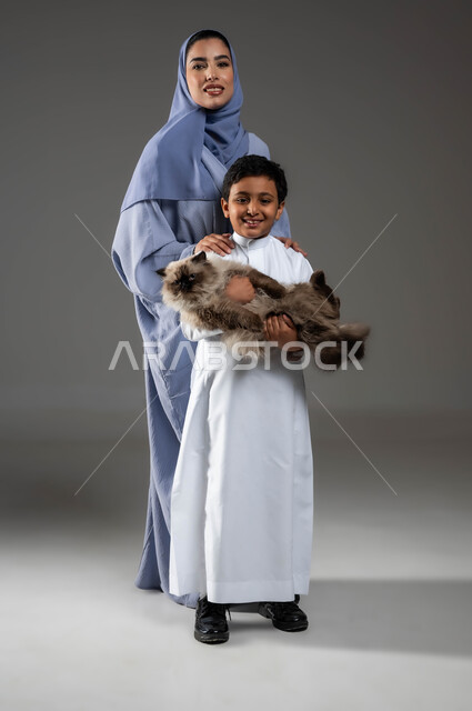 النظر الى الكاميرا بتعابير السعادة والسرور لاقتناء الحيوانات الأليفة، الأثر الايجابي لتربية القطط في المنازل، بورتريه لفتى عربي خليجي سعودي مبتسم يحمل قطة رمادية مدللة يقف بجانب والدته، مفهوم الرفق بالحيوان والعطب عليه، صورة الجسم بالطول الكامل، خلفية رمادية