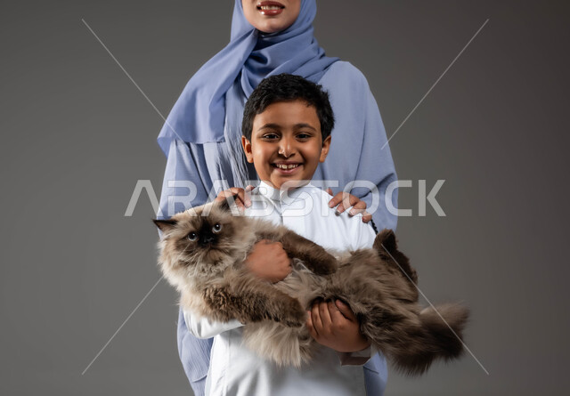 مفهوم الرفق بالحيوان والعطب عليه، النظر الى الكاميرا بتعابير السعادة والسرور لاقتناء الحيوانات الأليفة، الأثر الايجابي لتربية القطط في المنازل، بورتريه مقرب لفتى عربي خليجي سعودي مبتسم يحمل قطة رمادية مدللة يقف بجانب والدته، خلفية رمادية