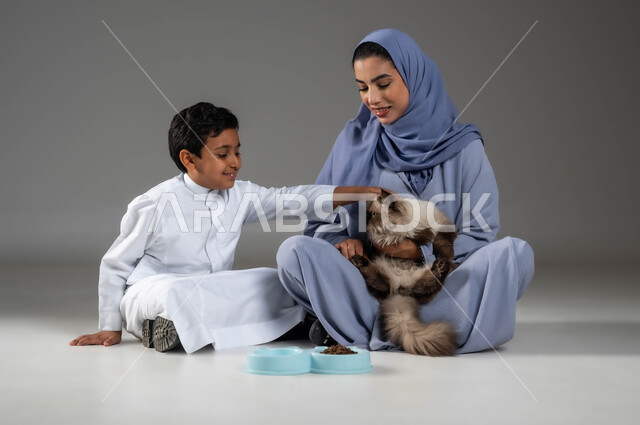 قضاء أوقات ممتعة في اللعب مع القط، بورتريه لأم عربية خليجية سعودية مبتسمة ترتدي العباءة والحجاب تجلس بجانب إبنها تقوم بإطعام القطة المدللة، رعاية وتربية القطط في المنازل، الاهتمام بصحة الحيوانات الأليفة، تقديم الرعاية الصحية والغذاء الجيد للقطط، خلفية رمادية