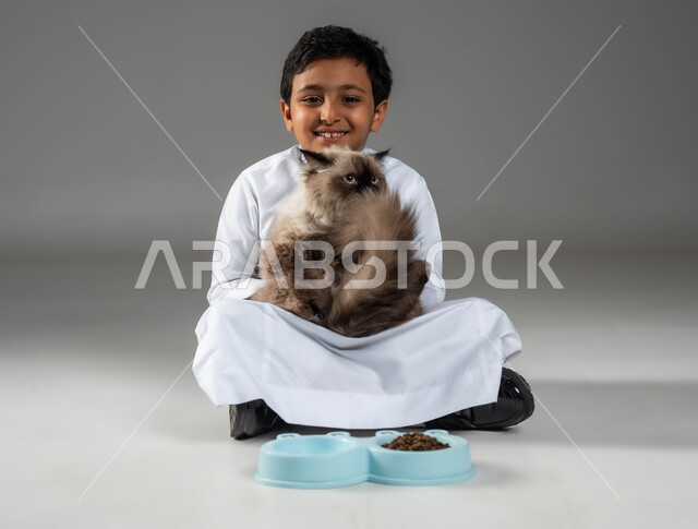النظر الى الكاميرا بتعابير السرور، توفير الرعاية الصحية والغذاء الجيد للقط، بورتريه لفتى عربي خليجي سعودي مبتسم يقوم بحمل القطة المدللة، مفهوم الرفق والاعتناء بالحيوان، الأثر الايجابي لتربية القطط في المنزل، الاهتمام بصحة الحيوانات الأليفة، خلفية رمادية
