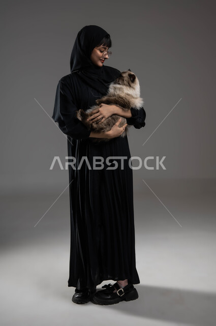 العطف والحنان على الحيوانات الأليفة، الاحساس بالسعادة أثناء قضاء وقت ممتع في اللعب مع القط، الأثر الايجابي لاقتناء الحيوانات المنزلية وتربيتها، بورتريه لشابة عربية خليجية سعودية مبتسمة ترتدي الحجاب والعباءة تقوم بحمل قطة رمادية، صورة الجسم بالكامل، خلفية رمادية