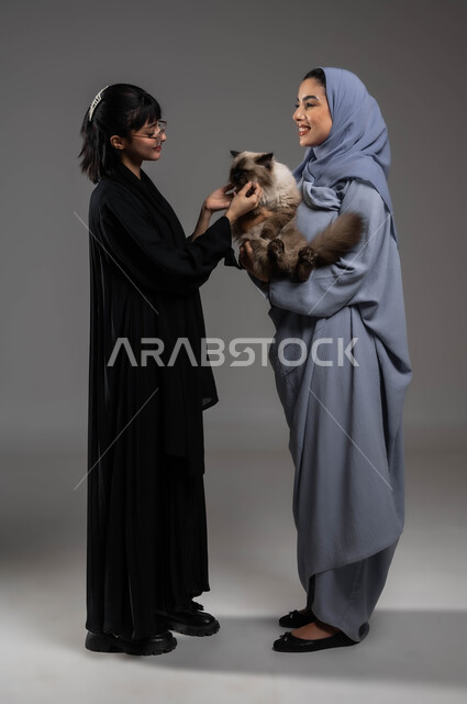 تحسين المزاج وملء الفراغ عند تربية الحيوان الأليف، مفهوم الرفق بالحيوان والعطف عليه، إيماءات السرور والسعادة، بورتريه لصديقتين عربيتين خليجيتين سعوديتين تستمتعان باللعب مع القطة الأليفة، صورة لكامل الجسم، خلفية رمادية