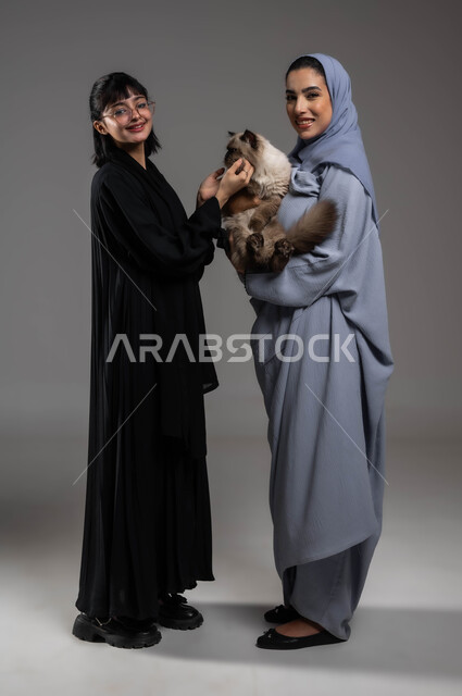 مفهوم الرفق بالحيوان والعطف عليه، النظر إلى الكاميرا بإيماءات السرور والسعادة، تحسين المزاج وملء الفراغ عند تربية الحيوان الأليف، بورتريه لصديقتين عربيتين خليجيتين سعوديتين تستمتعان باللعب مع القطة الأليفة، صورة لكامل الجسم، خلفية رمادية