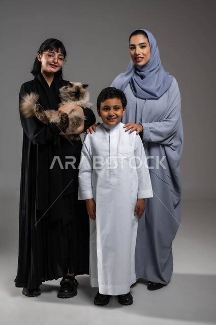 تحسين المزاج أثناء الاعتناء بالقطط داخل المنازل، الرفق بالحيوان والعطف عليه، فوائد تربية الحيوانات الأليفة للأطفال، النظر إلى الكاميرا بإيماءات السرور، بورتريه لعائلة عربية خليجية سعودية يقضون أوقات ممتعة مع القطة، صورة لكامل الجسم، خلفية رمادية