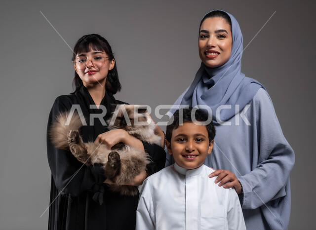 الاستمتاع بتربية الحيوان والاعتناء به، اهتمام العائلة بتربية أسماك الزينة والقطط الأليفة في المنزل، التسلية وتحسين المزاج باللعب مع القطة والسمكة، بورتريه مقرب لإمرأتين وولد عرب خليجيين سعوديين يجتمعان في البيت مع الحيوانات اللطيفة، خلفية رمادية