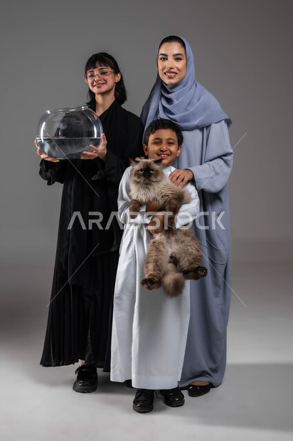 The family’s interest in raising ornamental fish and pet cats at home, having fun and improving the mood by playing with the cat and the fish, a portrait of two Saudi Gulf Arab women and a boy meeting at home with cute animals, the joy of raising an animal and taking care of it, full-length body portrait, gray background
