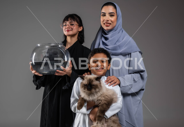 الأثر الإيجابي لتربية الحيوانات الأليفة على الأطفال، الرفق بالحيوان والعطف عليه، النظر إلى الكاميرا بإيماءات السرور، الاعتناء بالأسماك داخل المنازل، بورتريه مقرب لعائلة عربية خليجية سعودية يقضون أوقات ممتعة مع القطط، خلفية رمادية