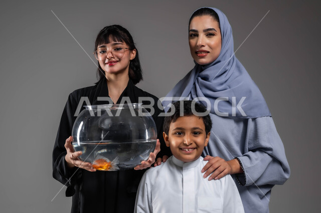 الاعتناء بالأسماك داخل المنازل، الرفق بالحيوان والعطف عليه، النظر إلى الكاميرا بإيماءات السعادة والسرور، بورتريه مقرب لعائلة عربية خليجية سعودية يقضون أوقات ممتعة مع الحيوانات الأليفة، خلفية رمادية