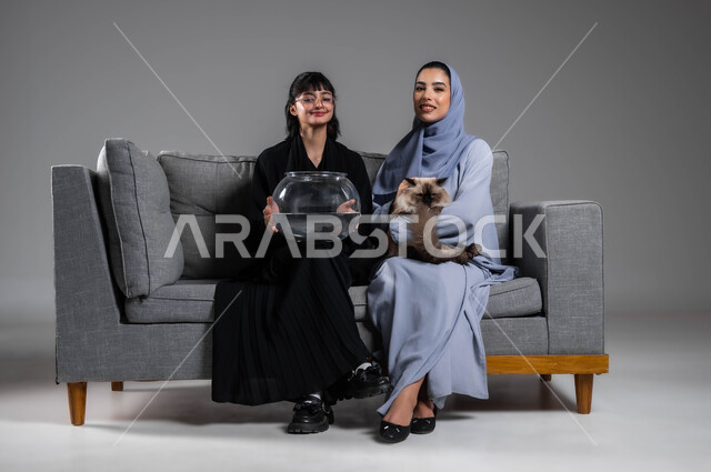 التسلية وتحسين المزاج باللعب مع القطة والسمكة، بورتريه لصديقتين عربيتين خليجيتين سعوديتين تجتمعان في البيت مع الحيوانات اللطيفة، اهتمام الفتيات بتربية أسماك الزينة والقطط الأليفة في المنزل،  متعة تربية الحيوان والاعتناء به، خلفية رمادية