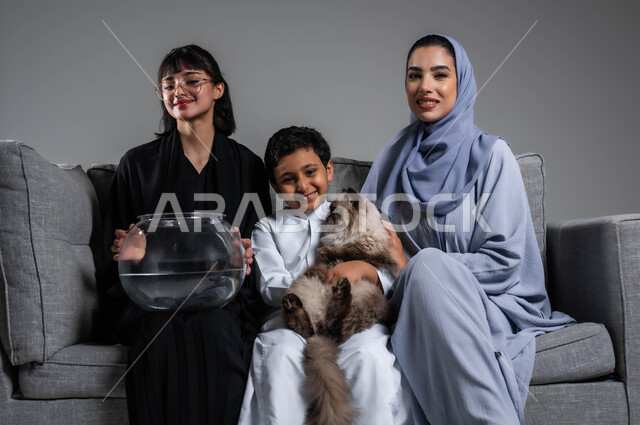 الاهتمام بأسماك الزينة الملونة، بورتريه لإمرأتين وولد عرب سعوديين خليجيين يقضون وقتا ممتعا مع الحيوانات الأليفة، تعليم الأبناء مفهوم الرفق بالحيوان والعطف عليه، تعابير المتعة باللعب واللهو مع القط الرمادي، خلفية رمادية