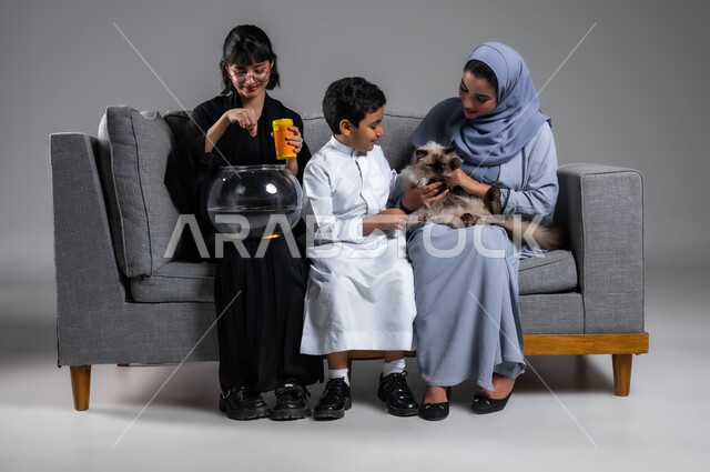 تعابير المتعة باللعب واللهو مع القط الرمادي، الاهتمام بأسماك الزينة وإطعامها، بورتريه لإمرأتين وولد عرب سعوديين خليجيين يقضون وقتا ممتعا مع الحيوانات الأليفة، تعليم الأبناء مفهوم الرفق بالحيوان والعطف عليه، خلفية رمادية