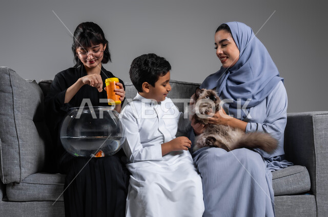 الاهتمام بأسماك الزينة وإطعامها، بورتريه لإمرأتين وولد عرب سعوديين خليجيين يقضون وقتا ممتعا مع الحيوانات الأليفة، تعليم الأبناء مفهوم الرفق بالحيوان والعطف عليه، الاستمتاع باللعب واللهو مع القط الرمادي، خلفية رمادية