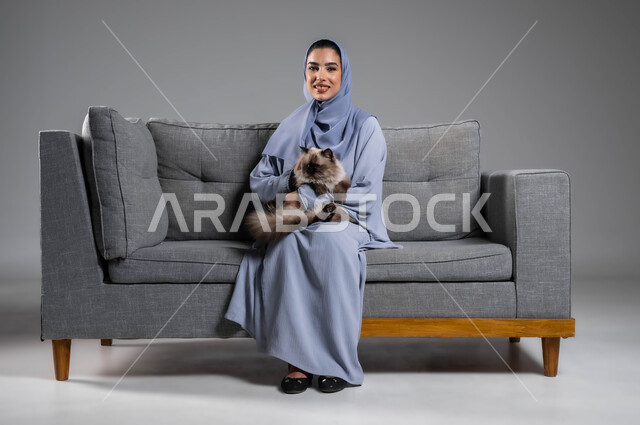 مفهوم الرفق بالحيوان والعطف عليه، الأثر الايجابي لاقتناء الحيوانات المنزلية وتربيتها، بورتريه لإمرأة عربية خليجية سعودية مبتسمة ترتدي الحجاب والعباءة تجلس على الاريكة تقوم بحمل قطة رمادية أليفة، الشعور بالسعادة أثناء قضاء وقت ممتع في اللعب مع القط، خلفية رمادية