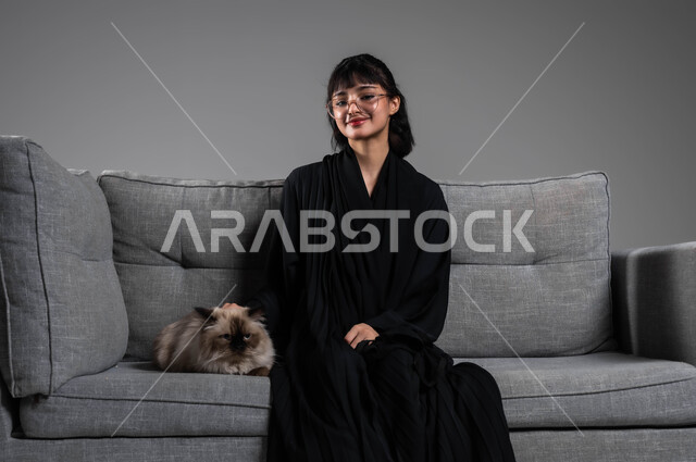 الرفق بالحيوان والعطف عليه، رعاية وتربية القطط في المنازل، بورتريه لشابة عربية خليجية سعودية مبتسمة ترتدي العباءة السوداء تجلس على الاريكة بجانب قطة رمادية أليفة، قضاء أوقات ممتعة في اللعب مع القط، الأثر الايجابي لاقتناء الحيوانات الأليفة، خلفية رمادية