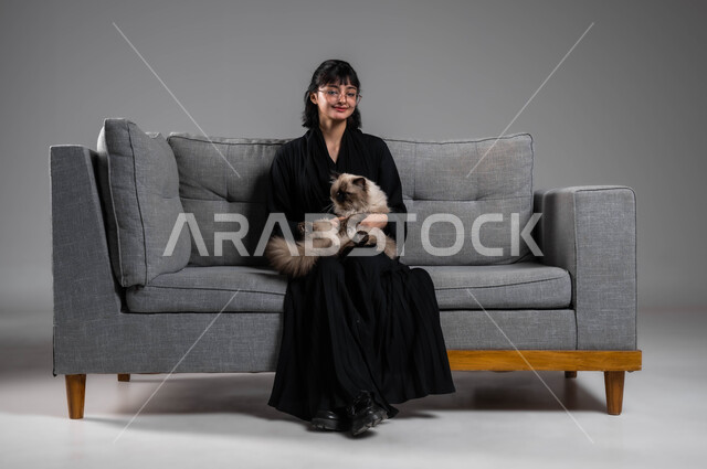 الرفق بالحيوان والعطف عليه، رعاية وتربية القطط في المنازل، بورتريه لشابة عربية خليجية سعودية مبتسمة ترتدي العباءة السوداء تجلس على الاريكة تقوم بمداعبة قطة رمادية أليفة، قضاء أوقات ممتعة في اللعب مع القط، الأثر الايجابي لاقتناء الحيوانات الأليفة، خلفية رمادية