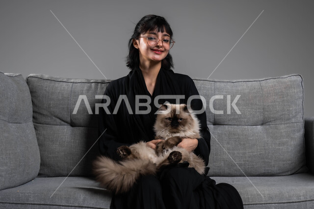 The positive impact of owning pets, kindness and compassion to animals, caring for and raising cats in homes, a portrait of a smiling Saudi Gulf Arab woman wearing a black abaya sitting on the sofa petting a pet gray cat, spending enjoyable times playing with the cat, gray background