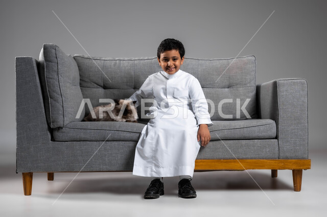 الشعور بالسعادة أثناء قضاء وقت ممتع في اللعب مع القط، الرفق بالحيوان والعطف عليه، الأثر الايجابي لاقتناء الحيوانات المنزلية وتربيتها، بورتريه لولد عربي خليجي سعودي مبتسم يرتدي الثوب التقليدي يجلس على الاريكة بجانب قطة رمادية أليفة، خلفية رمادية