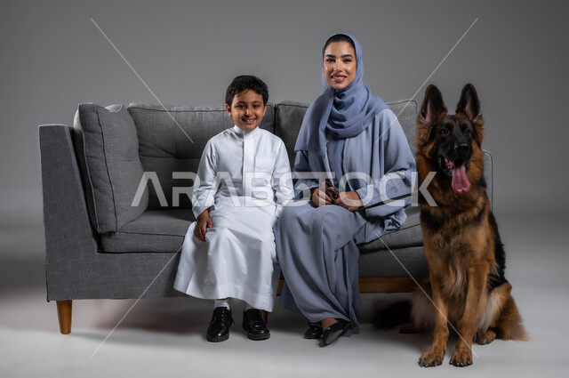 اللعب والمرح مع الحيوانات الأليفة، النظر إلى الكاميرا بإيماءات السرور، تربية الكلاب المنزلية، تعليم الأولاد العناية والرفق بالحيوان، بورتريه لأم عربية خليجية سعودية مبتسمة محجبة مع ابنها ترتدي العباءة تجلس على الاريكة بجانب كلب الهاسكي البني، خلفية رمادية