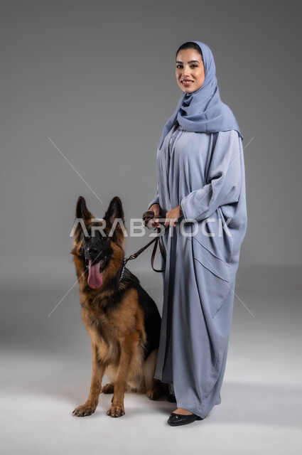 الاستمتاع بالتجول والمشي مع الكلاب، النظر الى الكاميرا بإيماءات السرور والسعادة، بورتريه لإمرأة عربية خليجية سعودية مبتسمة ترتدي العباءة والحجاب تقوم بمسك حبل المقود لكلب الهاسكي البني، اقتناء وتربية الحيوانات المنزلية والرفق بهم، صورة الجسم بالكامل، خلفية رمادية