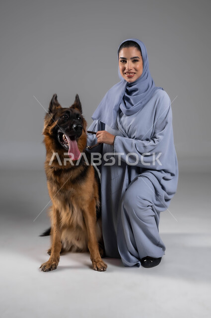 Acquiring and raising domestic dogs, playing and having fun with pets, a portrait of a smiling Saudi Arabian Gulf woman wearing an abaya and hijab sitting next to a large brown husky dog, the concept of animal welfare, looking at the camera with gestures of pleasure and happiness, gray background