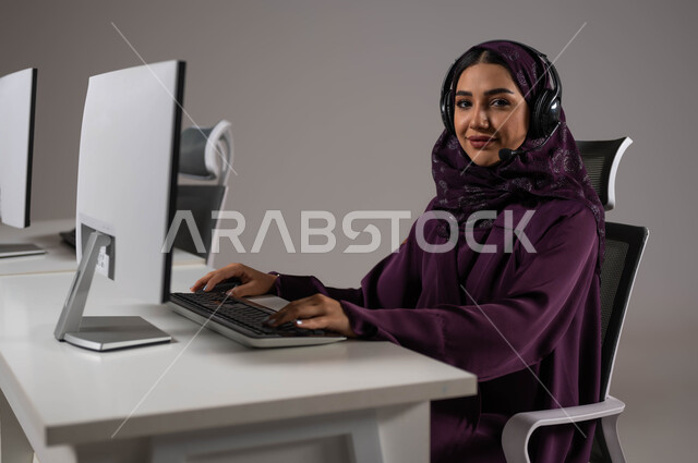 مفهوم الدعم الفني وخدمة العملاء, النظر إلى الكاميرا ،ارتداء سماعة الاتصال للإجابة عن الاستعلامات والاستفسارات ، استخدام التقنيات الحديثة، استقطاب و جذب المزيد من العملاء، بورتريه لسيدة عربيه سعوديه خليجيه مبتسمة محجبة تجلس في مقر العمل ,خلفية رمادية