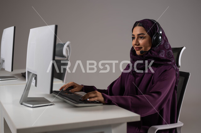 استقطاب و جذب المزيد من العملاء، مفهوم الدعم الفني وخدمة العملاء ،ارتداء سماعة الاتصال للإجابة عن الاستعلامات والاستفسارات ، استخدام التقنيات الحديثة، بورتريه لموظفة عربيه سعوديه خليجيه مبتسمة محجبة تجلس في مقر العمل ,خلفية رمادية