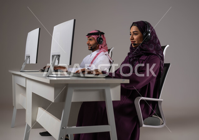 الجلوس في مقر الشركة، ايماءات الاندماج والتركيز، الإجابة على الاستفسارات و الاستعلامات، تقديم الخدمات المطلوبة والحرص على رضى العميل، ارتداء سماعة الاتصال للتواصل مع العملاء ،بورتريه لموظفان عربيان سعوديان خليجيان يعملان في خدمة العملاء ،خلفية رمادية