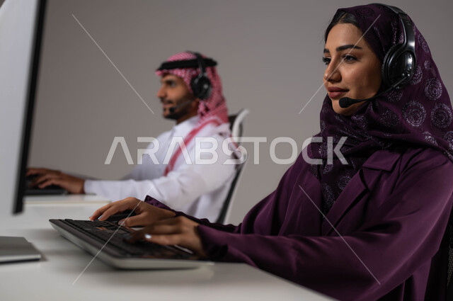 ايماءات الاندماج والتركيز، الإجابة على الاستفسارات و الاستعلامات، تقديم الخدمات المطلوبة والحرص على رضى العميل، ارتداء سماعة الاتصال للتواصل مع العملاء ،بورتريه مقرب لموظفان عربيان سعوديان خليجيان يعملان في خدمة العملاء ويجلسون في مقر العمل ،خلفية رمادية