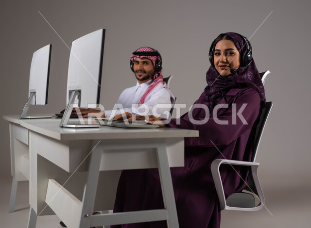 Looking at the camera, increasing the level of customer satisfaction, the concept of participation and cooperation at work, answering inquiries and enquiries, a portrait of a Saudi Gulf Arab man and woman working in customer service wearing a communication headset, Saudi professions and jobs, gray background