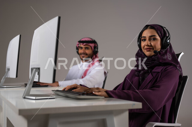 مفهوم المشاركة و التعاون في العمل ،الإجابة على الاستعلامات والاستفسارات, بورتريه مقرب لموظفان عربيان سعوديان خليجيان يعملان في خدمة العملاء ينظران إلى الكاميرا يرتديان سماعة الاتصال, زيادة مستوى رضا العميل، مهن ووظائف سعودية، خلفية رمادية