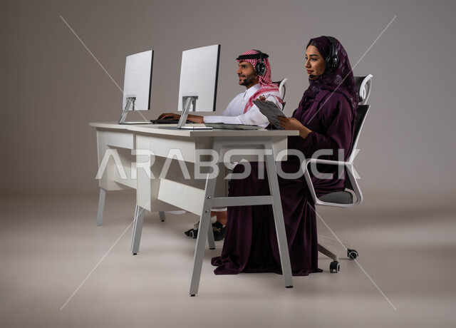 تدوين الشكاوي و الملاحظات، العمل في خدمة العملاء، متابعة وتطوير العمل في الشركات السعودية، ارتداء سماعة الاتصال للتواصل مع العميل ،الإجابة على الاستعلامات والاستفسارات, بورتريه لامرأة عربية سعودية خليجية مع زميلها يجلسون في مقر العمل ،خلفية رمادية