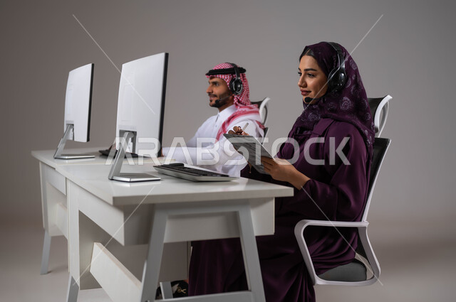 تسجيل و تدوين الشكاوي و الملاحظات، العمل في خدمة العملاء، متابعة وتطوير العمل في الشركات السعودية، ارتداء سماعة الاتصال للتواصل مع العميل ،الإجابة على الاستعلامات والاستفسارات, بورتريه لامرأة عربية سعودية خليجية مع زميلها يجلسون في مقر العمل ،خلفية رمادية