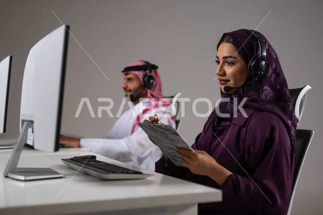 العمل في خدمة العملاء ،تدوين وتسجيل الملاحظات والشكاوي، متابعة وتطوير العمل في الشركات السعودية، ارتداء سماعة الاتصال للتواصل مع العميل ،الإجابة على الاستعلامات والاستفسارات, بورتريه مقرب لامرأة عربية سعودية خليجية مع زميلها يجلسون في مقر العمل ،خلفية رمادية