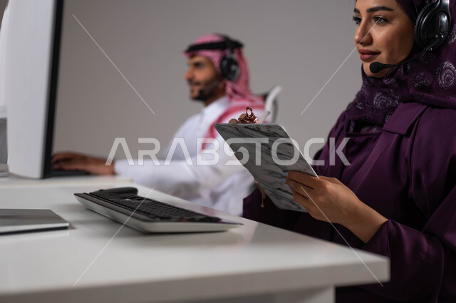 Taking and recording notes and complaints, following up and developing work in Saudi companies, working in customer service, wearing a communication headset to communicate with the customer, answering inquiries and inquiries, a close-up portrait of a Saudi Arabian Gulf woman with her colleague sitting in the workplace, gray background