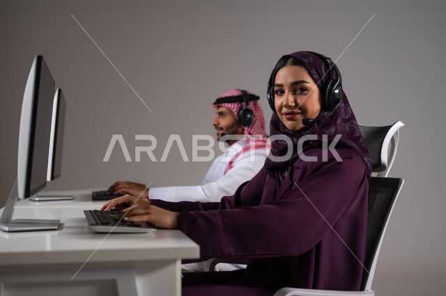 متابعة وتطوير العمل في الشركات السعودية ،مهن ووظائف سعودية، ارتداء سماعة الاتصال للتواصل مع العملاء ،الإجابة على الاستعلامات والاستفسارات, بورتريه مقرب لامرأة ورجل عربيان سعوديان خليجيان يعملان في خدمة العملاء ويجلسون في مقر العمل ،خلفية رمادية .