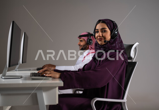 مهن ووظائف سعودية، ارتداء سماعة الاتصال للتواصل مع العملاء ،الإجابة على الاستعلامات والاستفسارات, بورتريه مقرب لامرأة ورجل عربيان سعوديان خليجيان يعملان في خدمة العملاء ويجلسون في مقر العمل ،متابعة وتطوير العمل في الشركات السعودية ،خلفية رمادية .