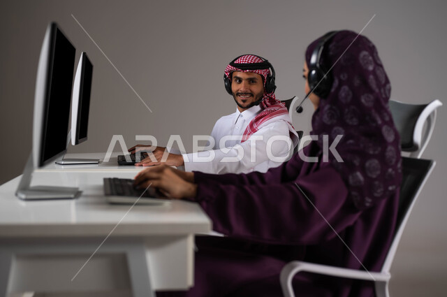 استخدام التقنيات الحديثة ،ارتداء سماعة الرأس والعمل على جهاز الحاسوب في خدمة العملاء، تعزيز مستوى رضا العميل، الإجابة عن الاستعلامات والاستفسارات, بورتريه لموظف عربي سعودي خليجي يجلس مع زميلته في مقر الشركة وينظران لبعضهما بإيماءات السرور، خلفية رمادية .