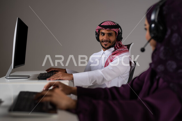 تعابير الثقة بالنفس، ارتداء سماعة الرأس والعمل على جهاز الحاسوب في خدمة العملاء، تعزيز مستوى رضا العميل، الإجابة عن الاستعلامات والاستفسارات, بورتريه لموظف عربي سعودي خليجي يجلس مع زميلته في مقر الشركة وينظران لبعضهما بإيماءات السرور، خلفية رمادية .