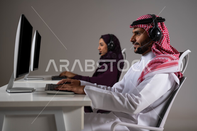 ارتداء سماعة الرأس والعمل على جهاز الحاسوب في خدمة العملاء، دمج التكنولوجيا و التقنيات الحديثة بالحياة العملية ،تعزيز مستوى رضا العميل، الإجابة عن الاستعلامات والاستفسارات, بورتريه لرجل و امرأة عربيان سعوديان خليجيان يجلسان في مقر الشركة، خلفية رمادية .