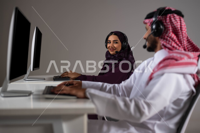 ارتداء سماعة الرأس والعمل على جهاز الحاسوب في خدمة العملاء، تعابير الثقة بالنفس، تعزيز مستوى رضا العميل، الإجابة عن الاستعلامات والاستفسارات, بورتريه لموظفان عربيان سعوديان خليجيان يجلسان في مقر الشركة وينظران لبعضهما بإيماءات السرور،  خلفية رمادية .