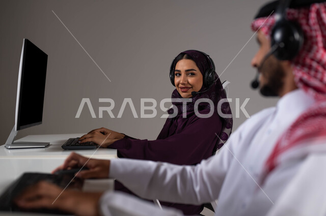 Expressions of self-confidence, enhancing the level of customer satisfaction, answering queries and enquiries, a portrait of two Saudi Arabian Gulf employees sitting at the company headquarters and looking at each other with pleased gestures, wearing a headset and working on the computer in customer service, gray background.