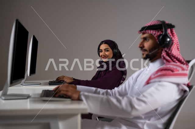 تعزيز مستوى رضا العميل، الإجابة عن الاستعلامات والاستفسارات,  تعابير و إيماءات الثقة بالنفس، بورتريه لموظفان عربيان سعوديان خليجيان يجلسان في مقر الشركة ،النظر إلى الكاميرا ،ارتداء سماعة الرأس والعمل على جهاز الحاسوب في خدمة العملاء، خلفية رمادية .