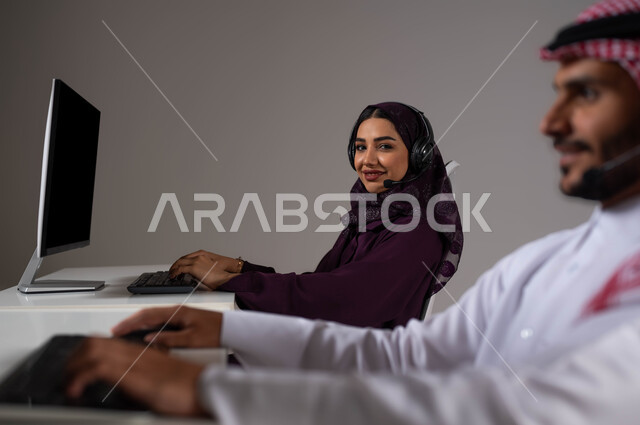 Gestures of self-confidence, enhancing the level of customer satisfaction, answering queries and enquiries, a portrait of two Saudi and Gulf Arab employees sitting at the company headquarters, looking at the camera, wearing a headset and working on the computer in customer service, gray background.