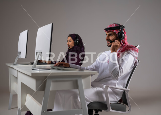 الإجابة على الاستفسارات و الاستعلامات ،وظائف ومهن سعودية، تقديم الخدمات المطلوبة والحرص على رضى العميل، ارتداء سماعة الاتصال للتواصل مع العملاء ،بورتريه لموظفان عربيان سعوديان خليجيان يعملان في خدمة العملاء ويجلسون في مقر العمل ،خلفية رمادية