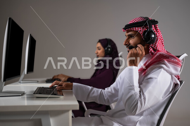 Providing the required services and ensuring customer satisfaction, answering inquiries and inquiries, wearing a communication headset to communicate with customers, a close-up portrait of two Saudi and Gulf Arab employees working in customer service and sitting in the workplace, Saudi professions and jobs, gray background