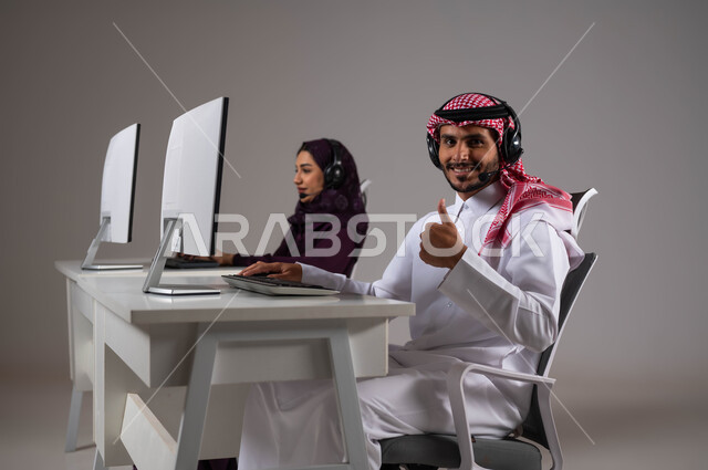 النظر إلى الكاميرا بايماءات السرور ورفع اليد بعلامة الجودة ،تطوير و متابعه العمل في الشركات السعودية، الإجابة على الاستفسارات و الاستعلامات, ارتداء سماعة الاتصال للتواصل مع العملاء ،بورتريه لموظفان عربيان سعوديان خليجيان يعملان في خدمة العملاء ،خلفية رمادية