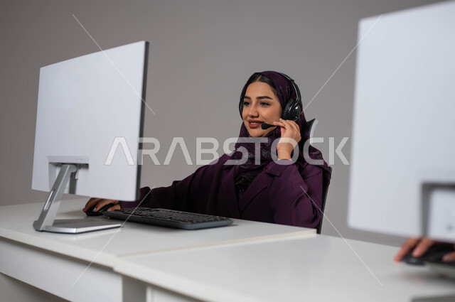انجاز الاعمال والمهام المكتبيه ،الإجابة على الأسئلة والاستفسارات, قسم الاستعلامات والاتصال، مفهوم خدمة العملاء، وظائف ومهن سعودية، بورتريه لموظفة عربية خليجية سعودية محجبة مبتسمة تجلس في مقر الشركة ترتدي سماعات الرأس ،خلفية رمادية